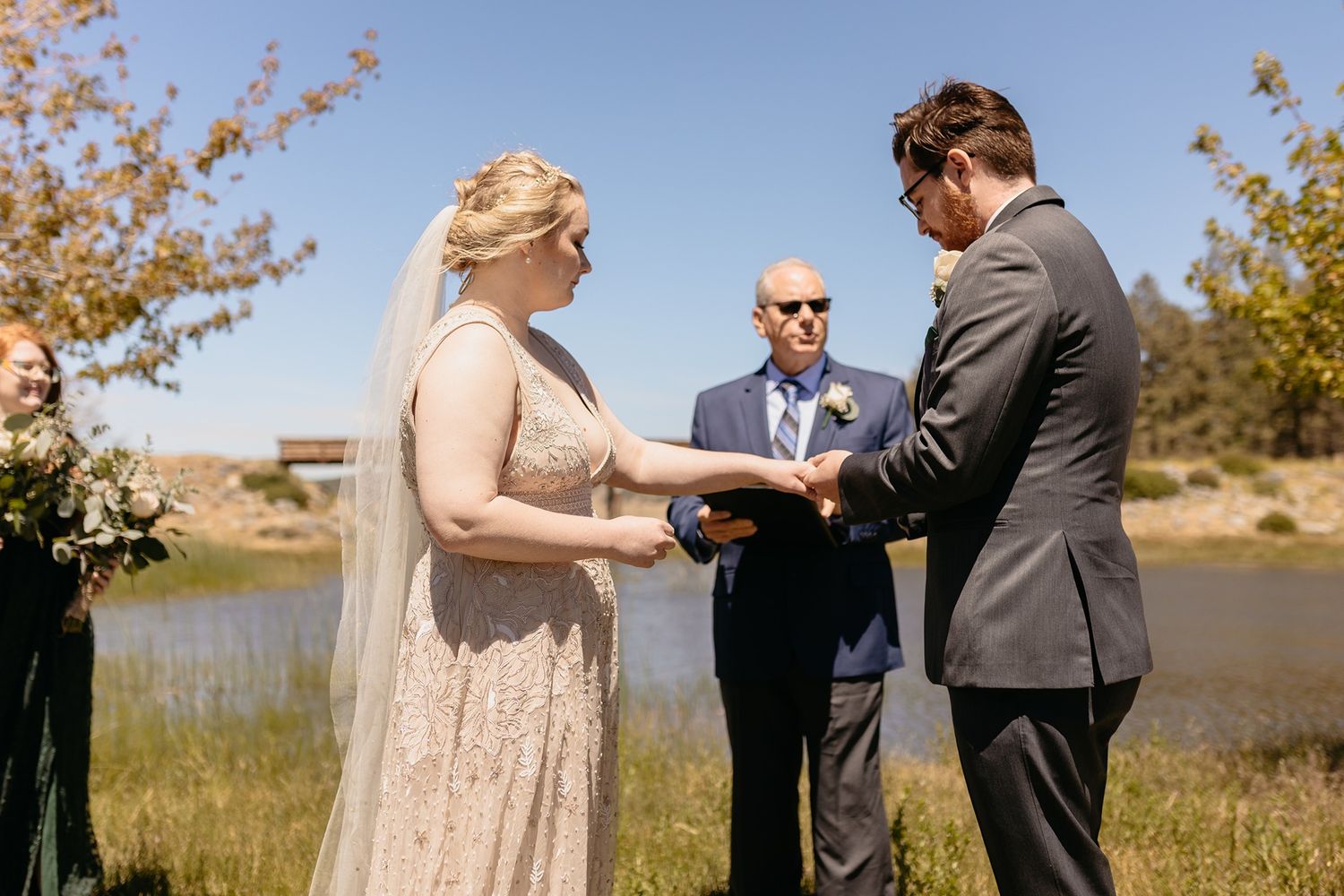 Outdoor wedding ceremony by a lake with bride in lace gown, groom in gray suit, and officiant under blue sky at Lake Cuyamaca, Julian.