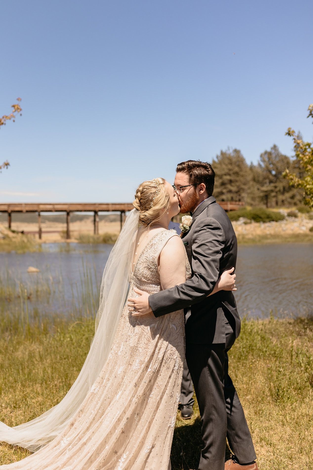 Bride and groom share a romantic kiss outdoors near a river, bride wearing a lace gown with veil on a sunny day at Lake Cuyamaca, Julian.