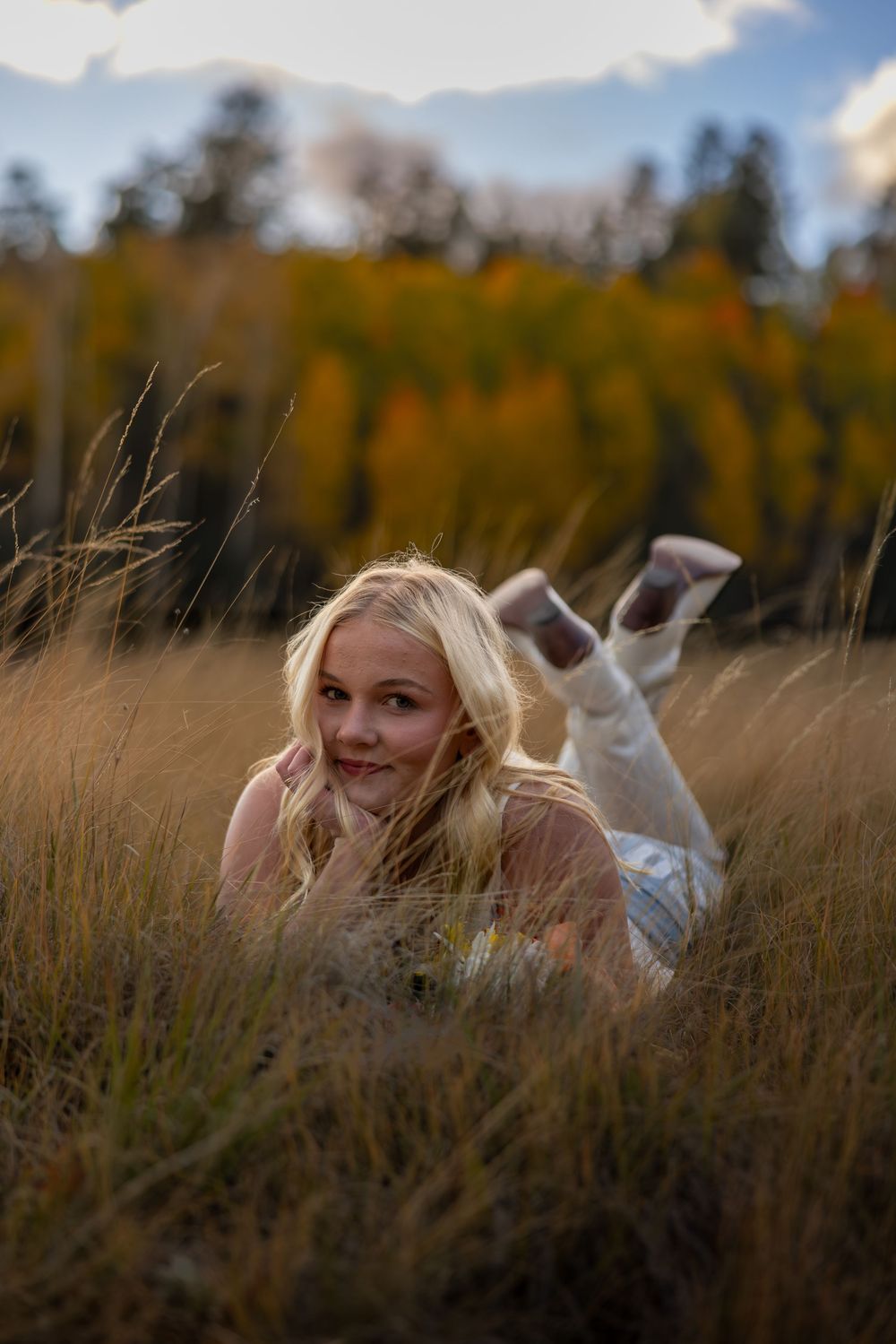 Blonde woman lies in tall golden grass field with autumn foliage in background on cloudy day.
