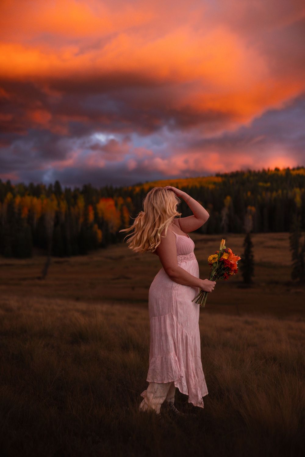 Woman in pink dress holding colorful bouquet in autumn field under dramatic red sunset sky.