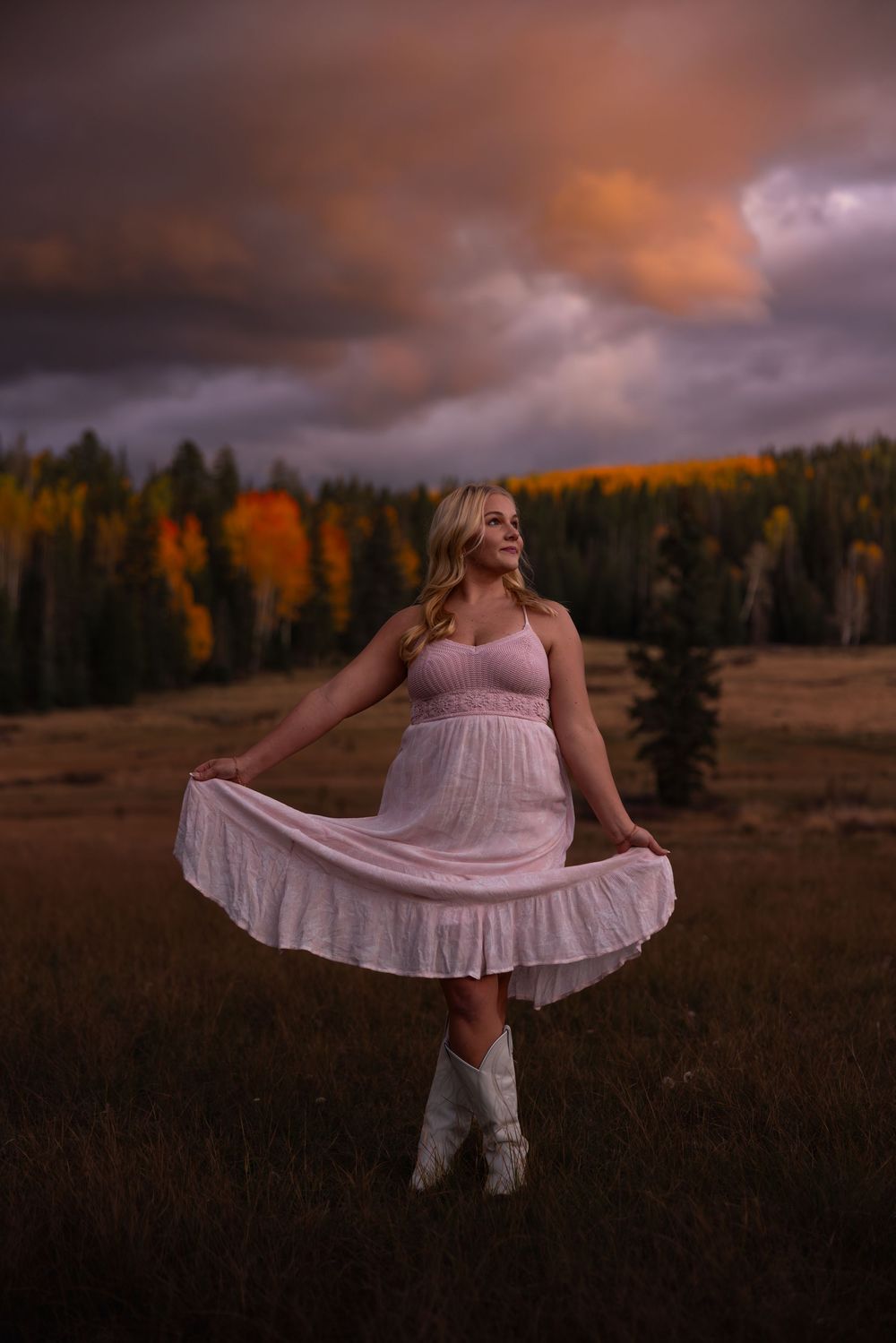 Blonde woman in white dress and cowboy boots twirling in autumn meadow under stormy sunset sky.
