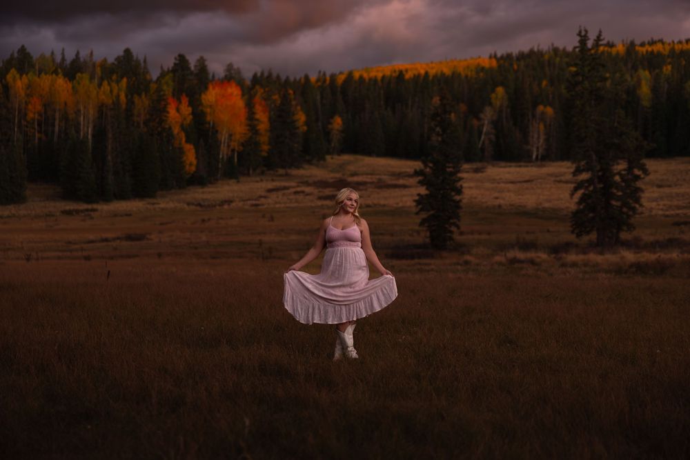 Woman in white dress spinning in dark autumn meadow with colorful fall trees at dusk.