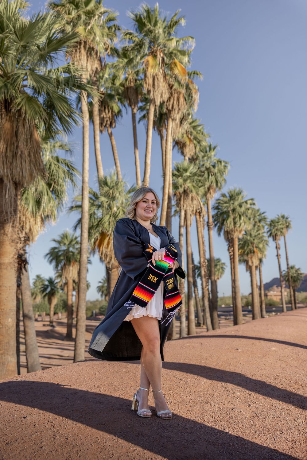 Graduate wearing cap and gown with colorful stole posing among tall palm trees in Arizona.