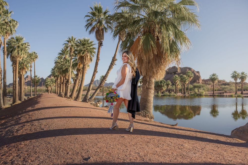 Woman in white dress carrying bouquet walking along palm-lined lakeside path in bright sunshine.