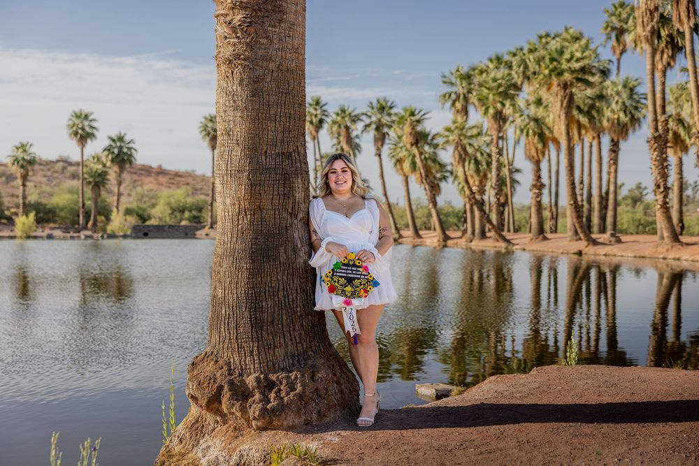 Woman in white dress leaning against palm tree by a reflective lake surrounded by palms.