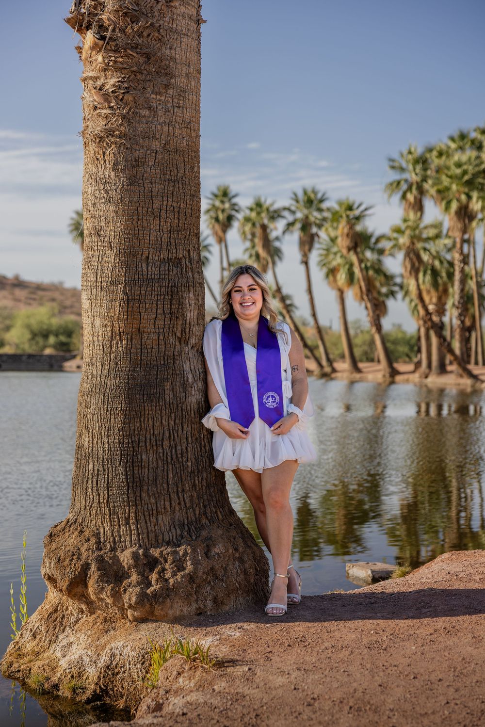 Graduate in white dress and purple BSN stole posing by palm tree at lakeside park.