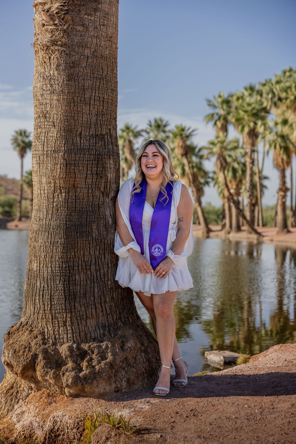 Smiling graduate in white dress with purple stole leaning against palm tree by lake.