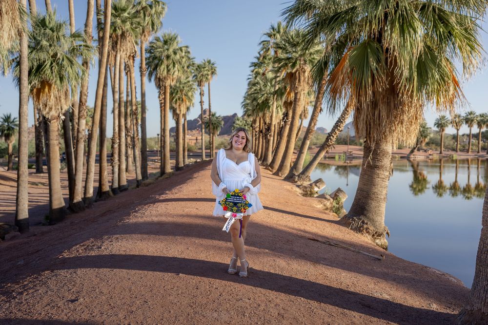 Bride in short white dress holding bouquet walking along palm-lined path near reflective lake.