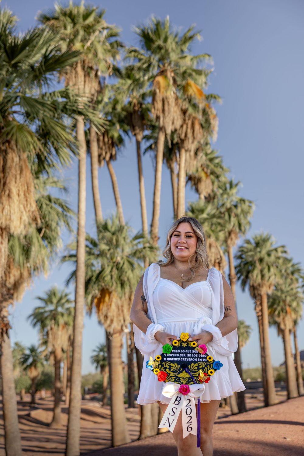 Graduate in white dress holding colorful cap and BSN 2026 sign among tall palm trees.