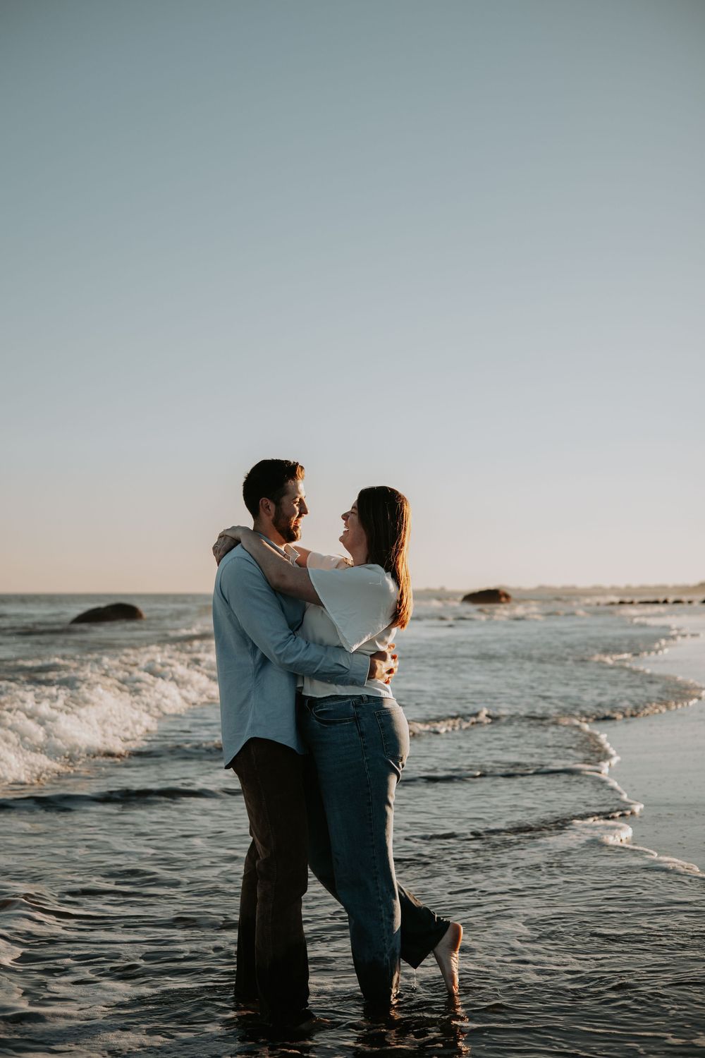 Couple embraces while standing in ocean waves at sunset as water splashes around them on the beach. Connecticut Wedding and Elopement.