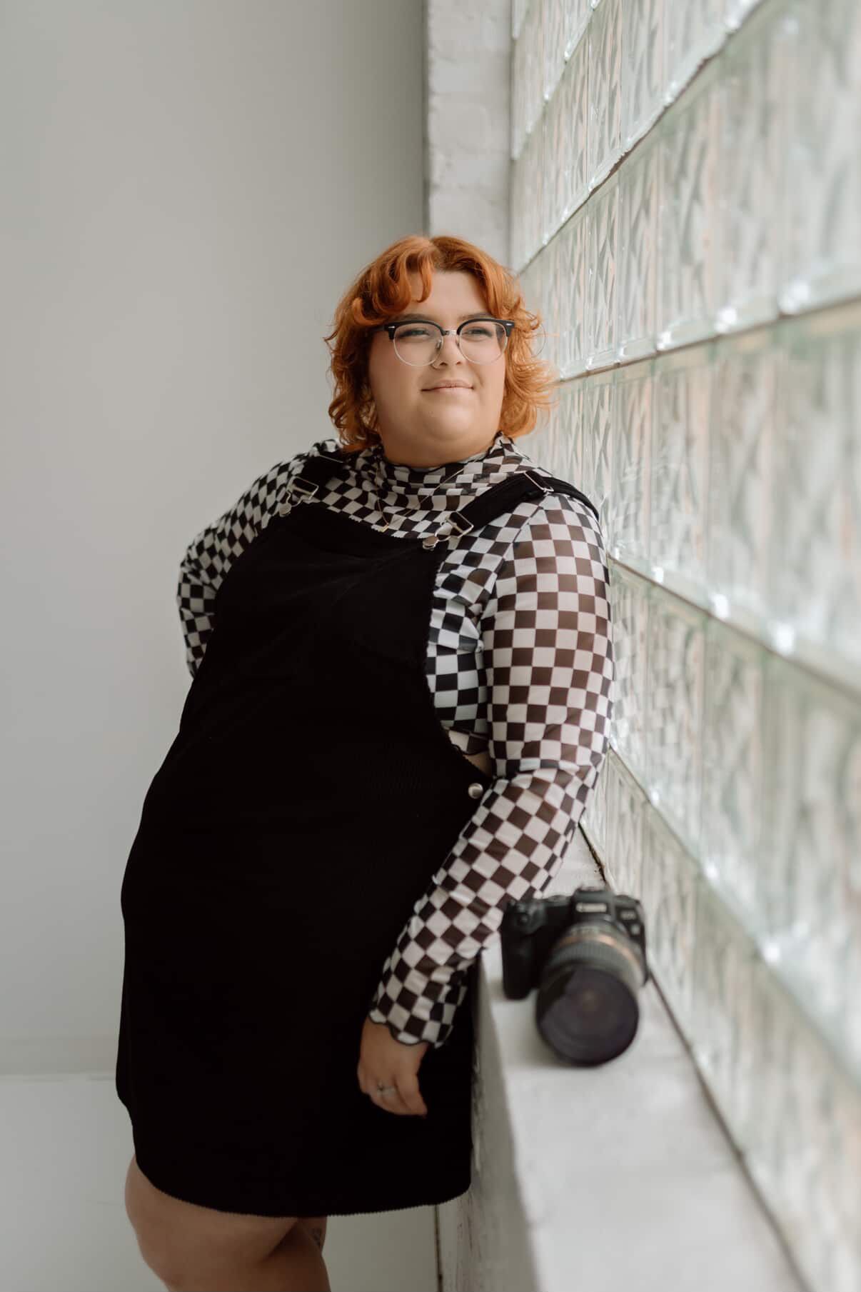 Person wearing floral dress and glasses poses with camera in indoor setting.