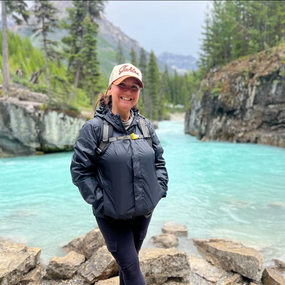 Person in dark jacket standing on rocky shore beside turquoise mountain lake.