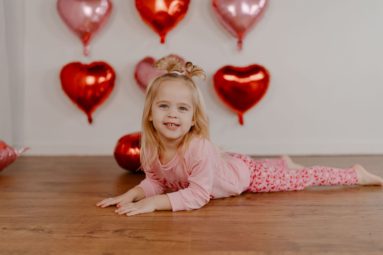 Red heart balloons decorate a backdrop as a young person in pink poses on a wooden floor.