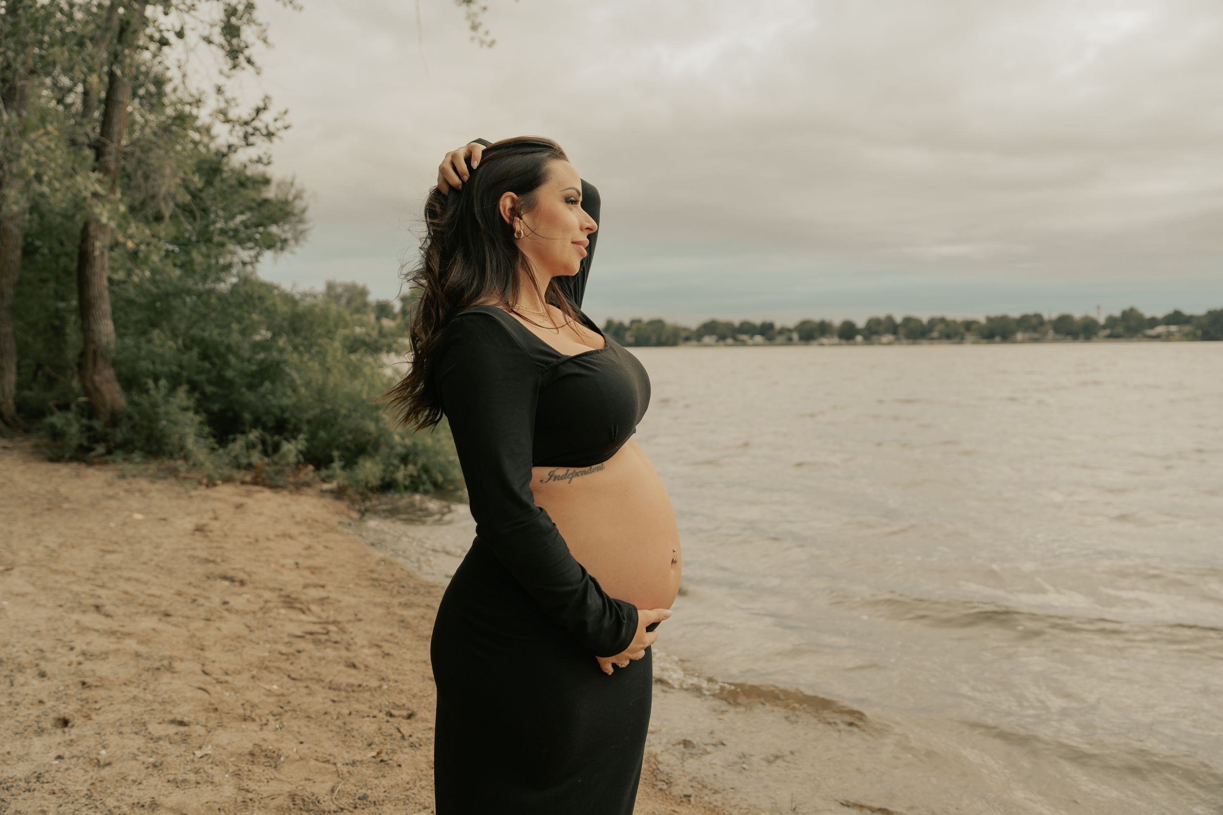An expectant mother poses beside a lake wearing a black dress.