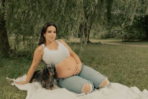 A pregnant woman relaxes on a blanket outdoors with a small dog.