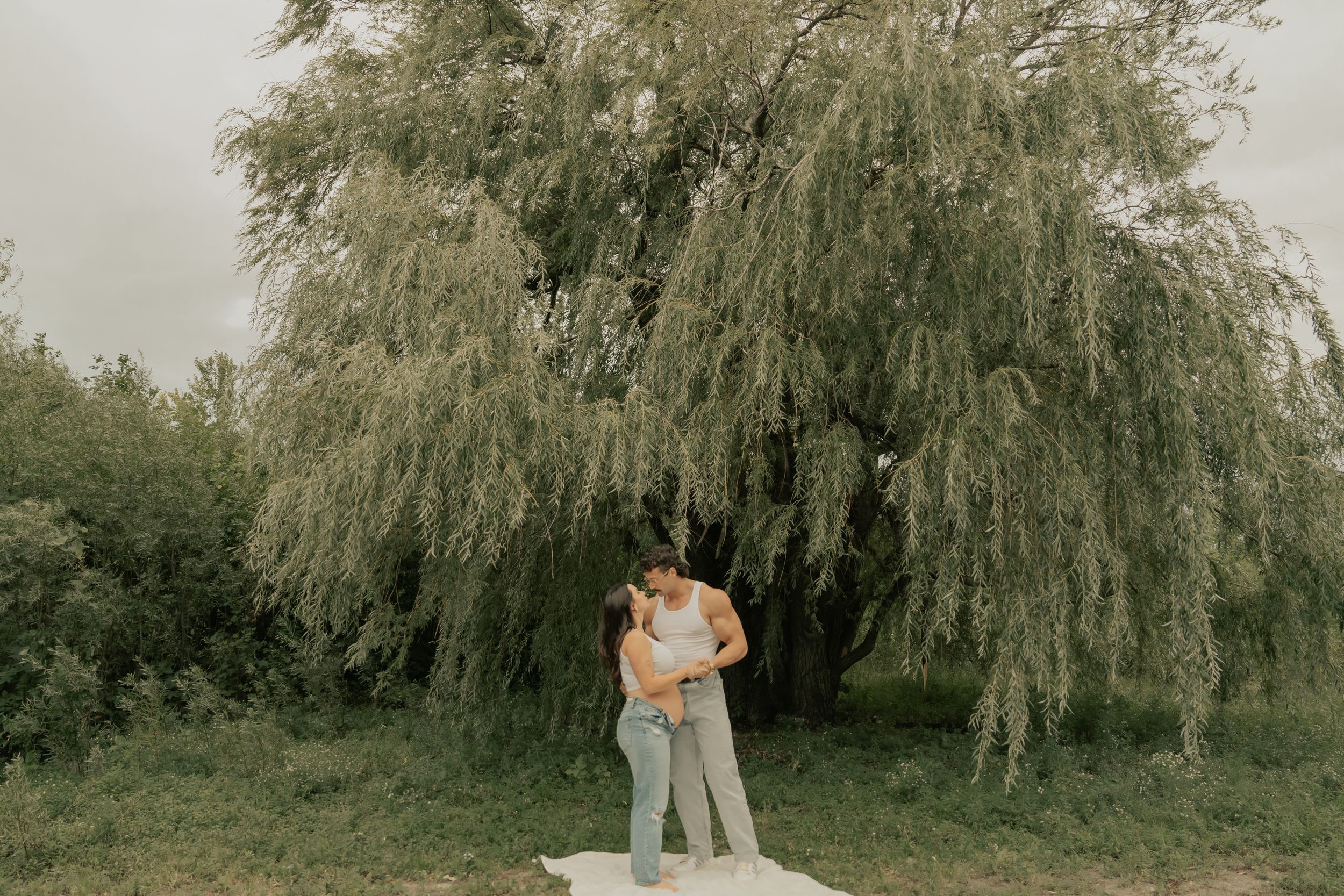 A couple standing together on a white blanket under a drooping willow tree.