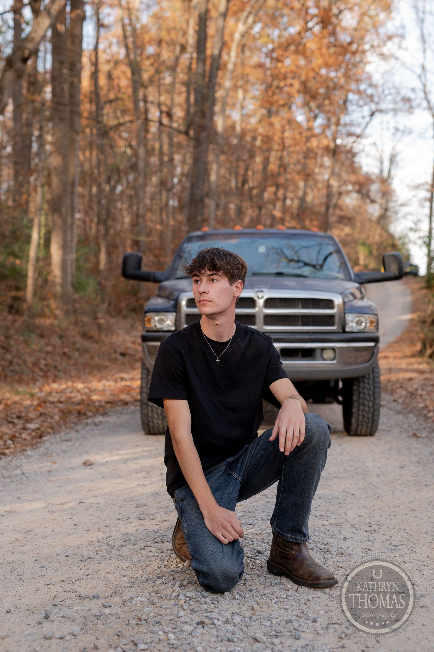 Person kneeling on gravel road in fall scenery with Dodge Ram truck parked against autumn forest backdrop.