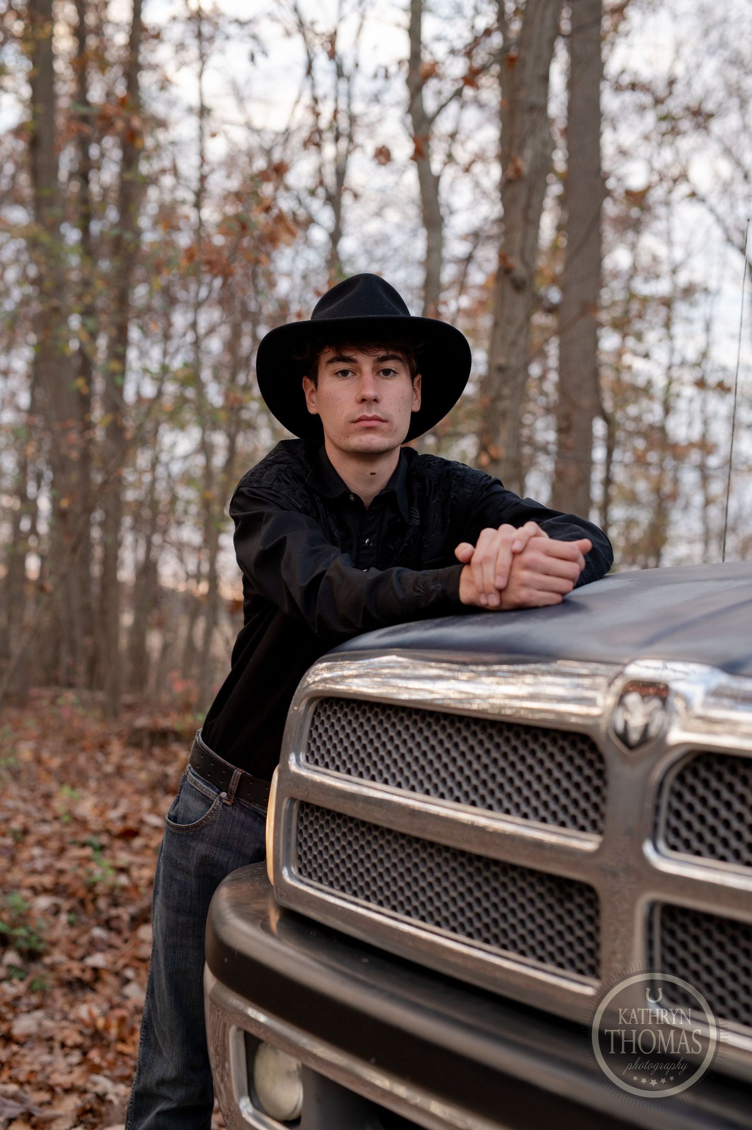 Someone in a black hat leans against a silver Dodge Ram truck in a wooded setting.