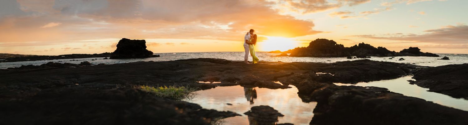 Couple silhouetted against vibrant sunset sky standing on rocky coastline with tide pool reflections below.