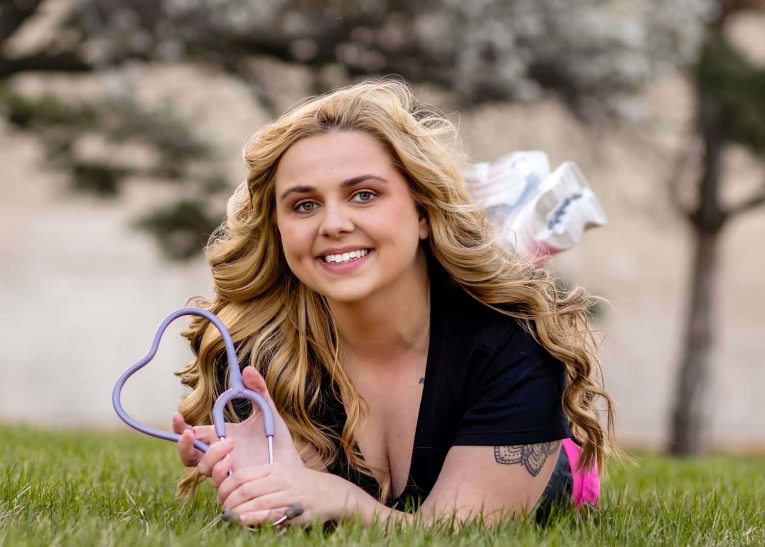 A blonde woman in a black top lies on green grass outdoors while smiling at the camera.