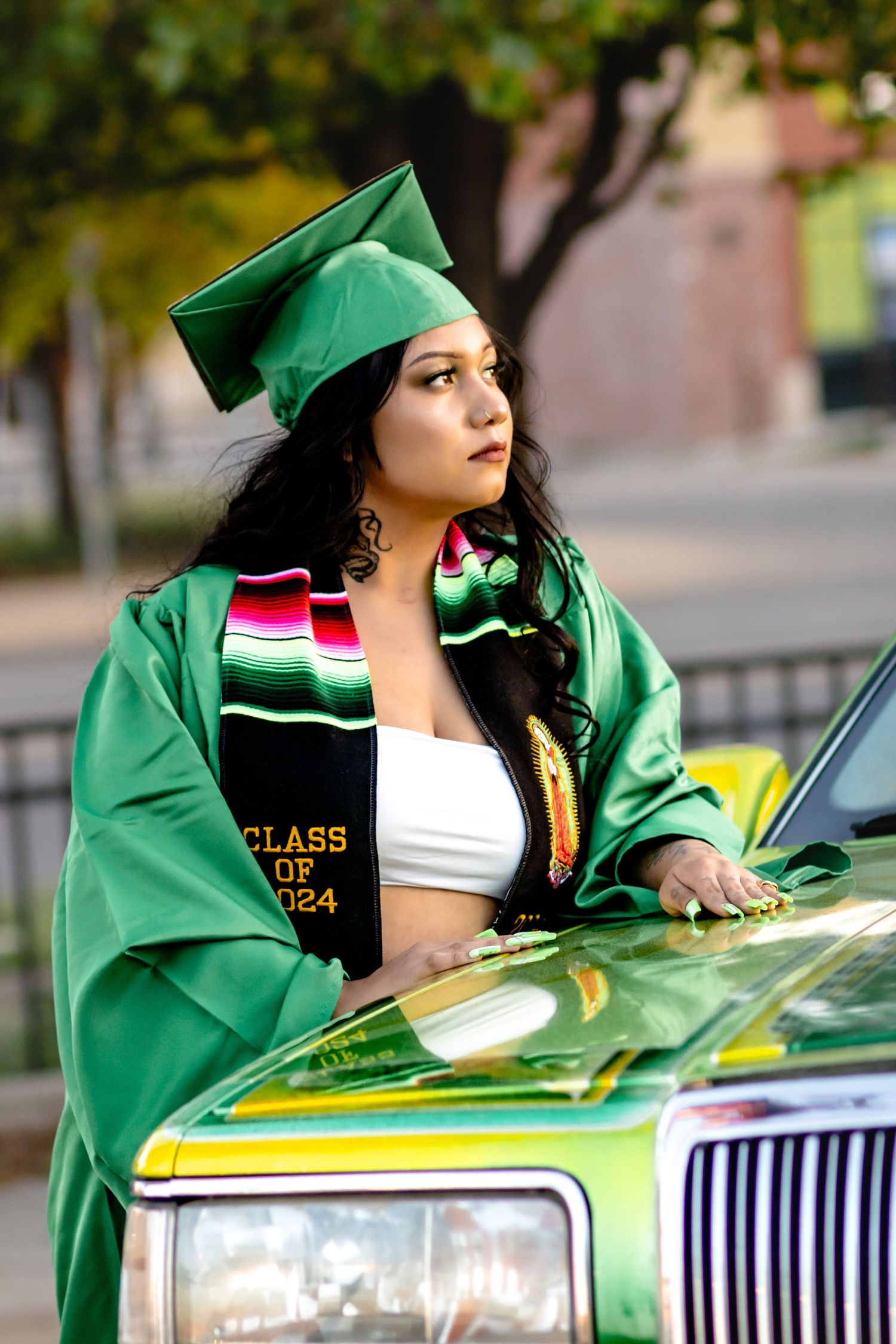 Graduate in emerald green cap and gown with cultural stole poses next to green car during graduation photoshoot.
