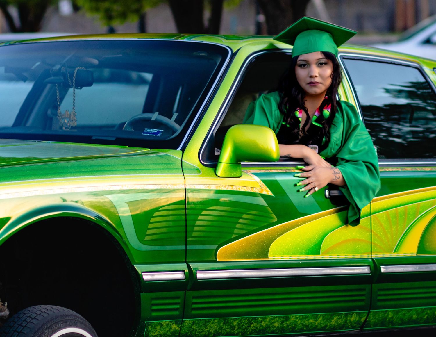 Graduate wearing green cap and gown poses by bright lime-green lowrider car.