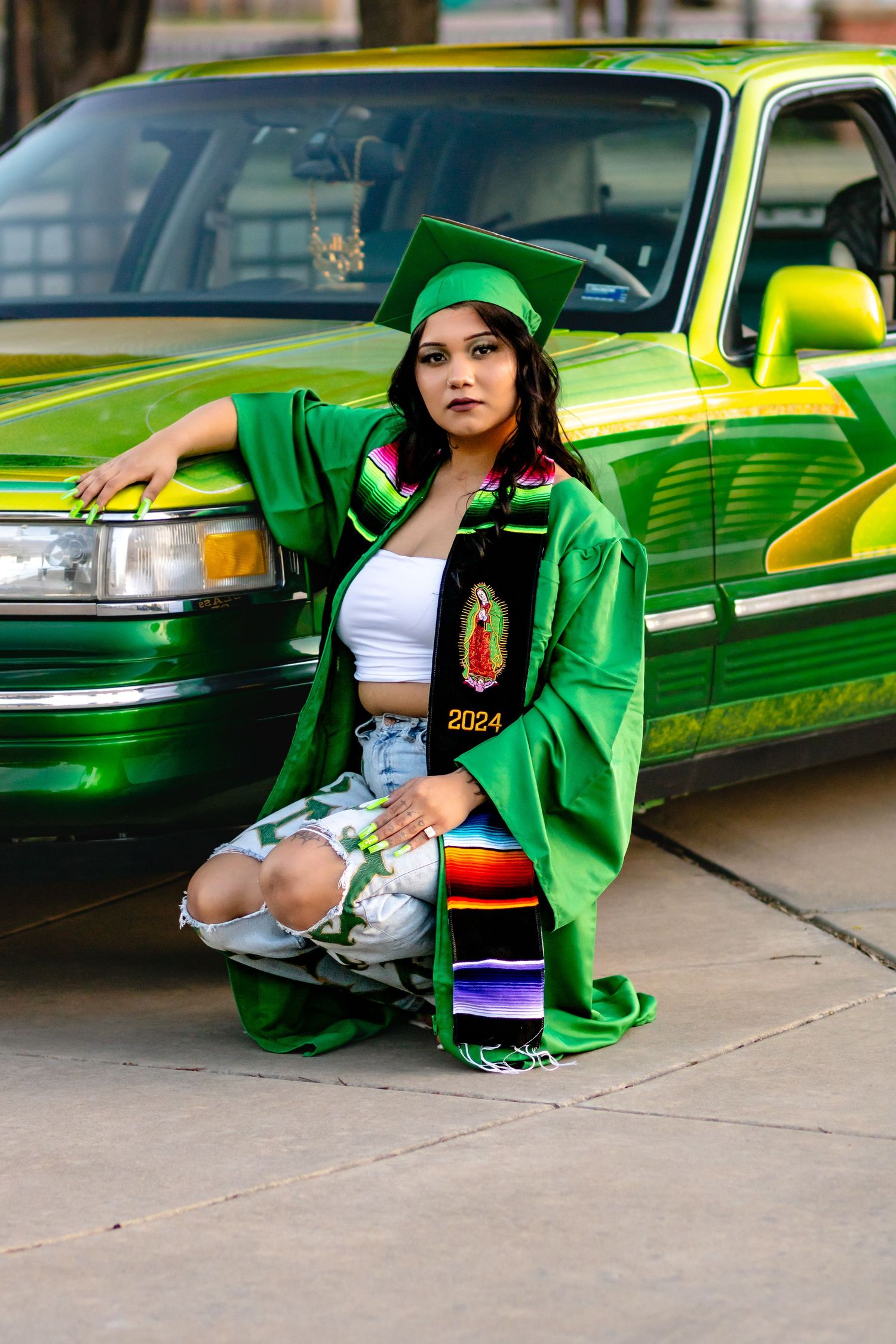 Graduate in green cap and gown poses next to metallic lime green lowrider car on sidewalk.