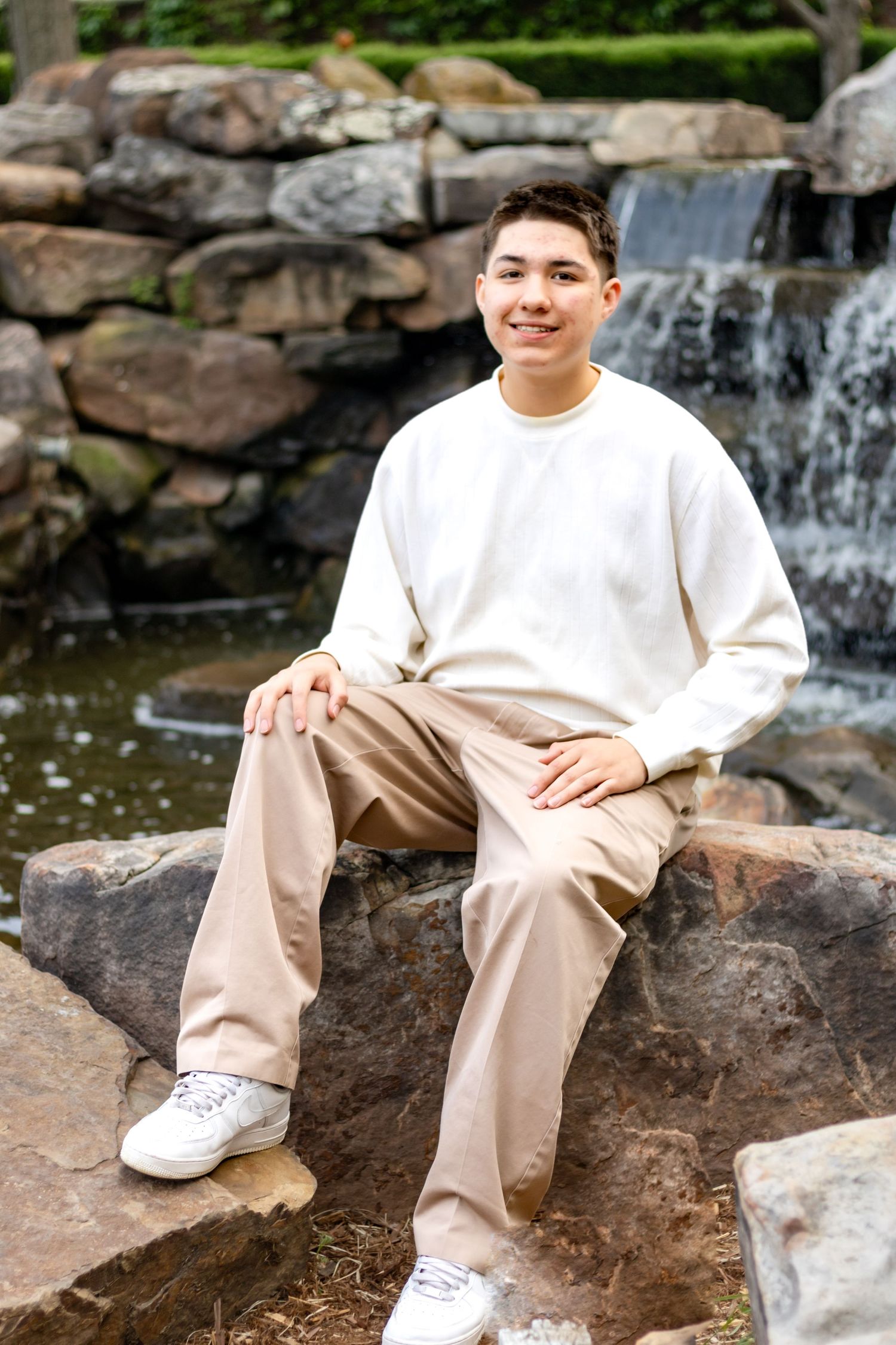 Someone in a white sweater and khaki pants sits on rocks by a cascading waterfall feature in a garden setting.