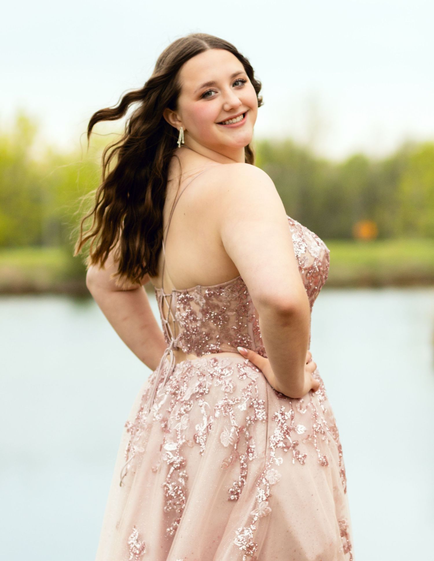 A person wearing a strapless pink formal gown with beaded details poses by a lake during an outdoor photo session.