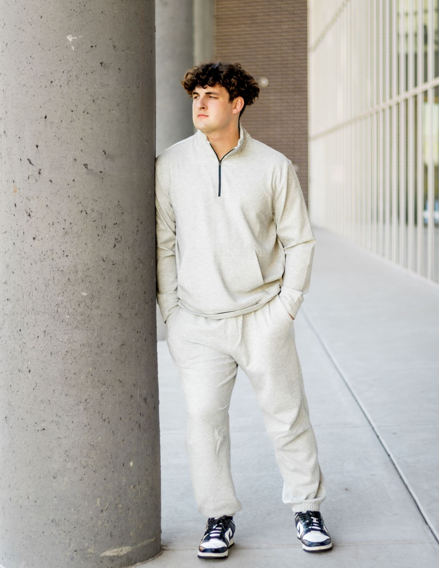 Person wearing all white casual outfit with quarter-zip sweater and sneakers poses against concrete pillar in hallway.