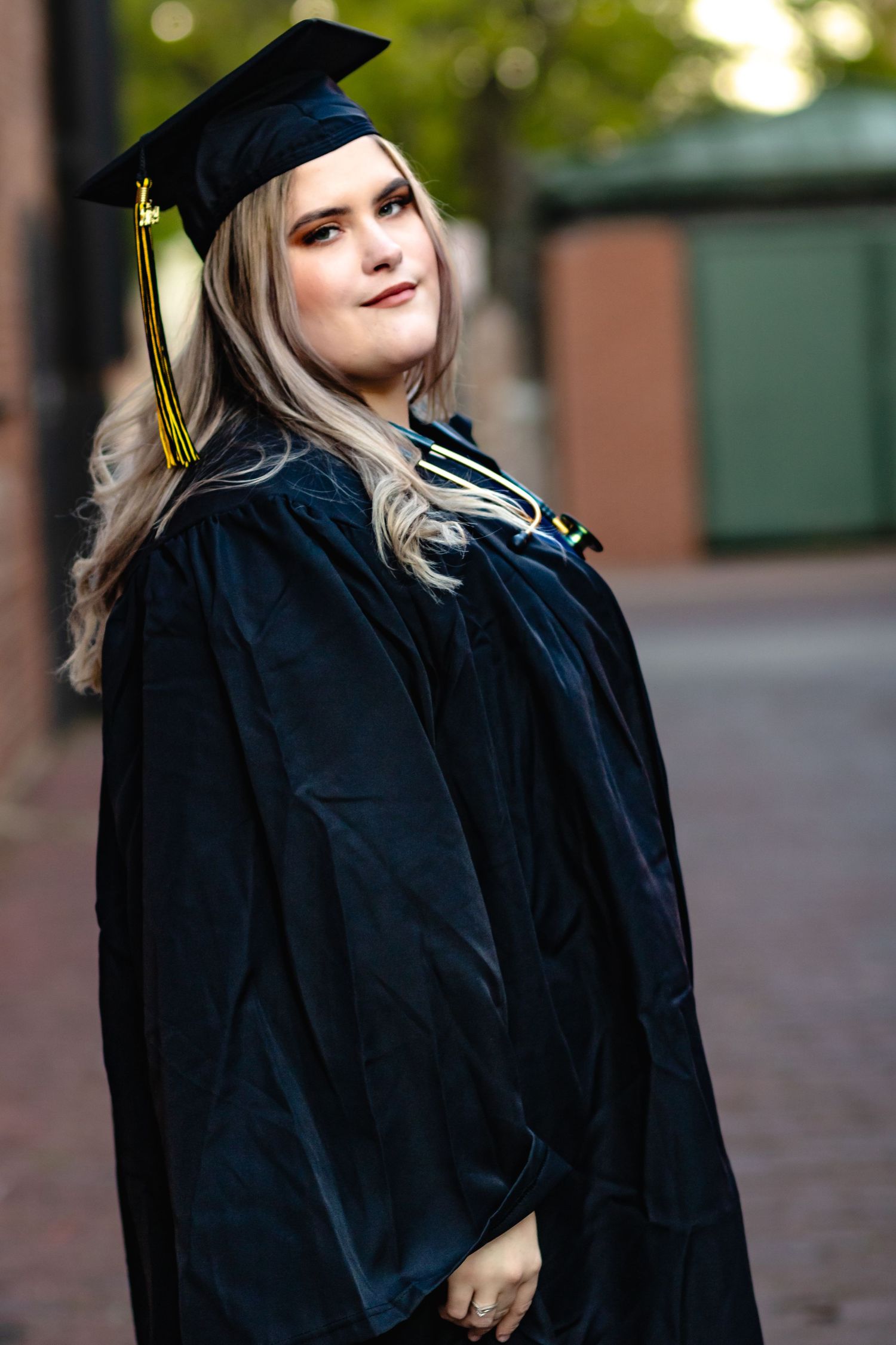 Graduate in traditional black cap and gown stands elegantly against blurred brick background.