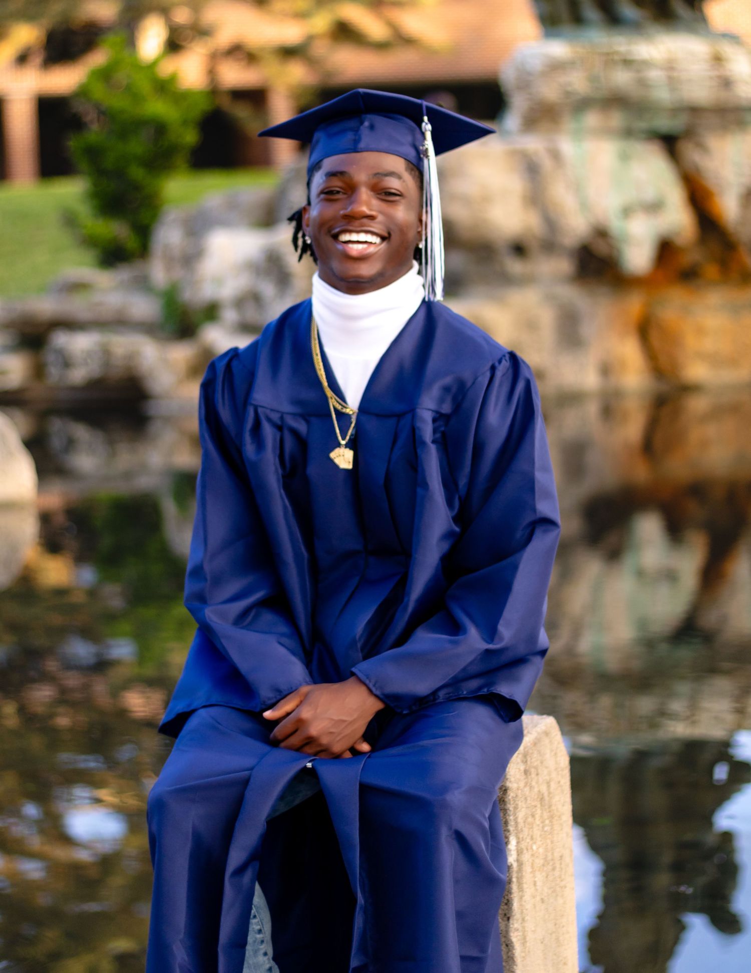 A graduate in navy blue cap and gown sits outdoors smiling brightly near a scenic rock wall and water feature.