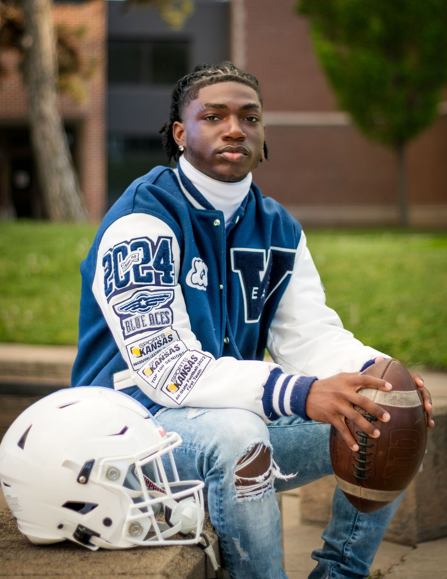 High school athlete in blue and white varsity letterman jacket poses outdoors with football and helmet on campus.
