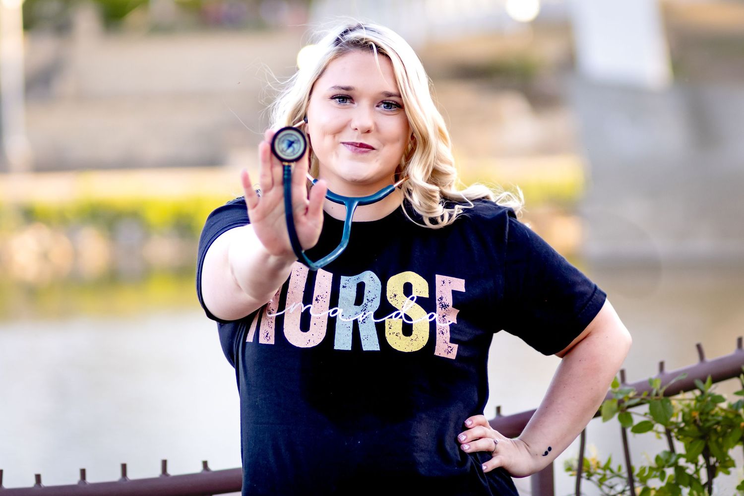 A healthcare professional in a navy 'NURSE' t-shirt holds up a stethoscope outdoors during a sunny day.