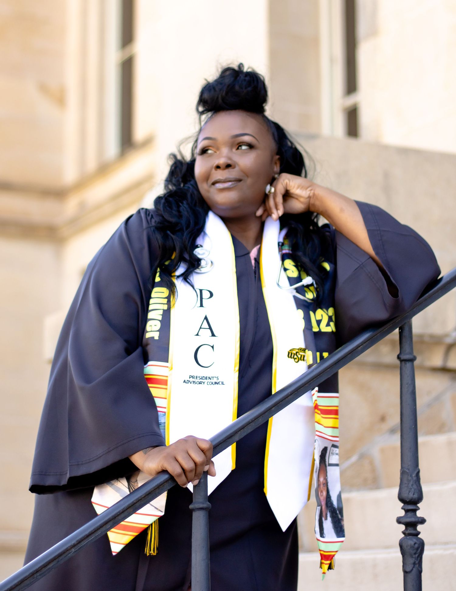 College graduate wearing graduation stole, cap and gown holding staff and smiling while posing outdoors.
