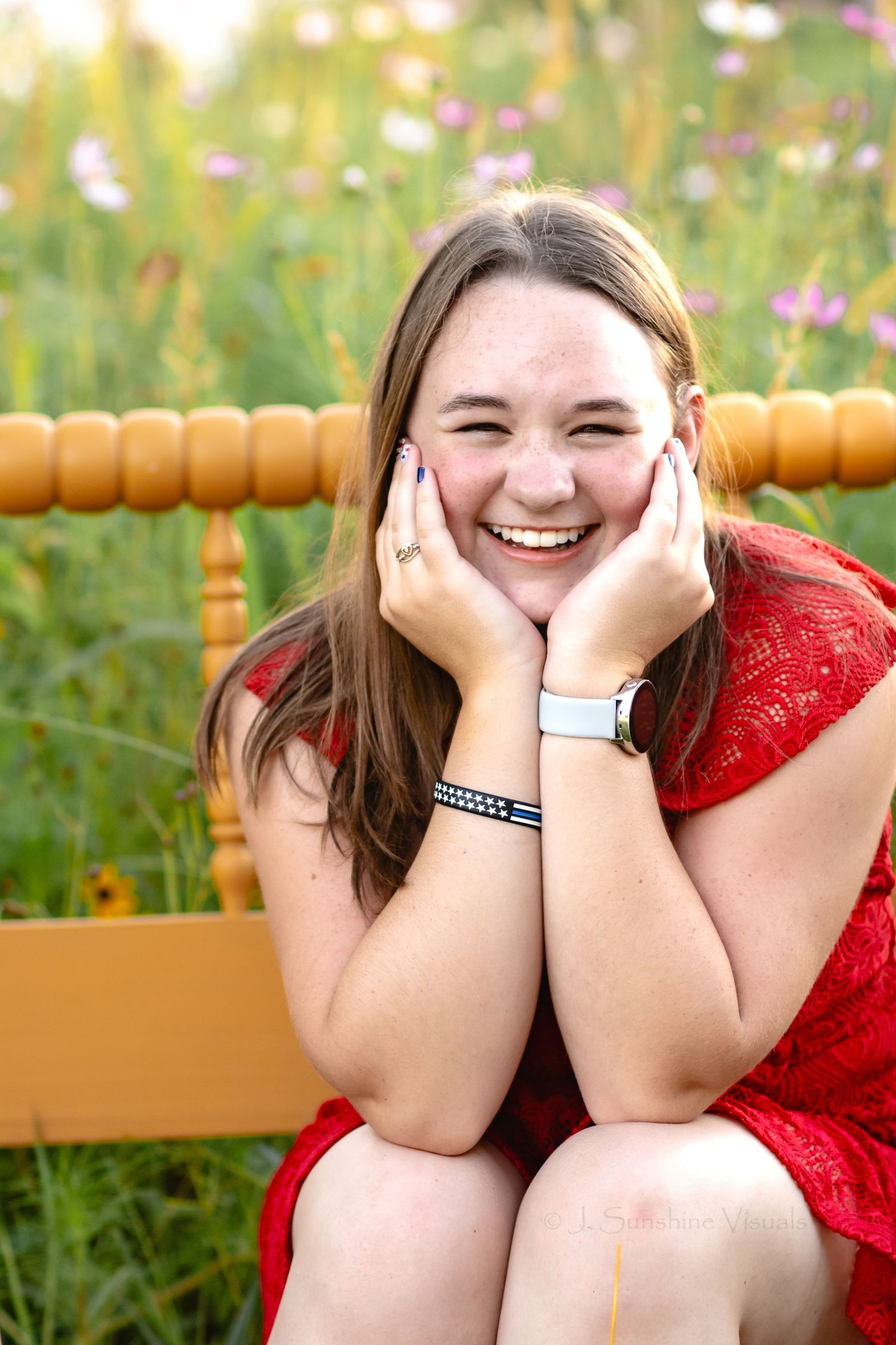 Someone wearing a red dress laughs joyfully while sitting in a wooden chair against a backdrop of wildflowers.