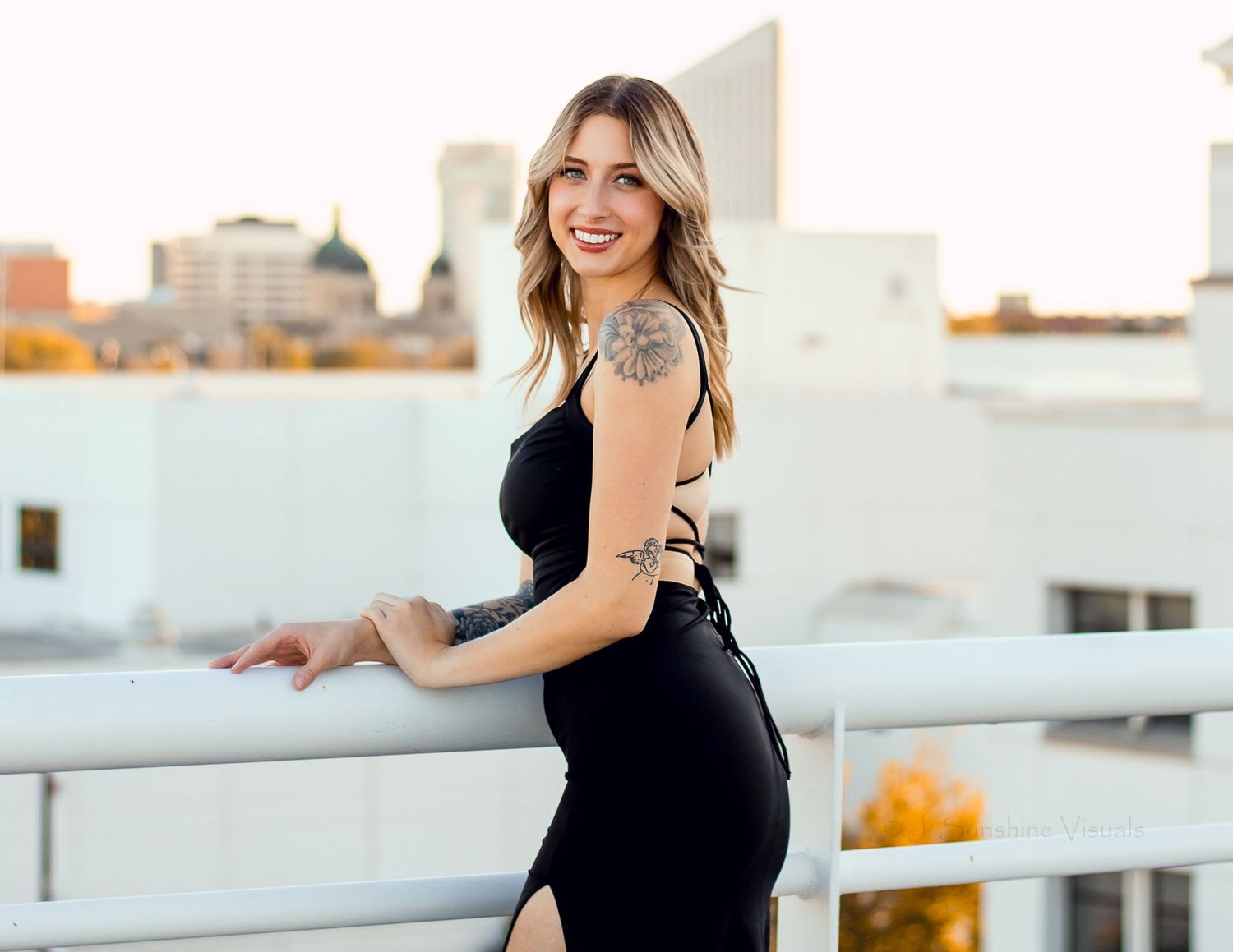 Woman in sleeveless black dress poses on city rooftop at sunset with urban skyline in background.