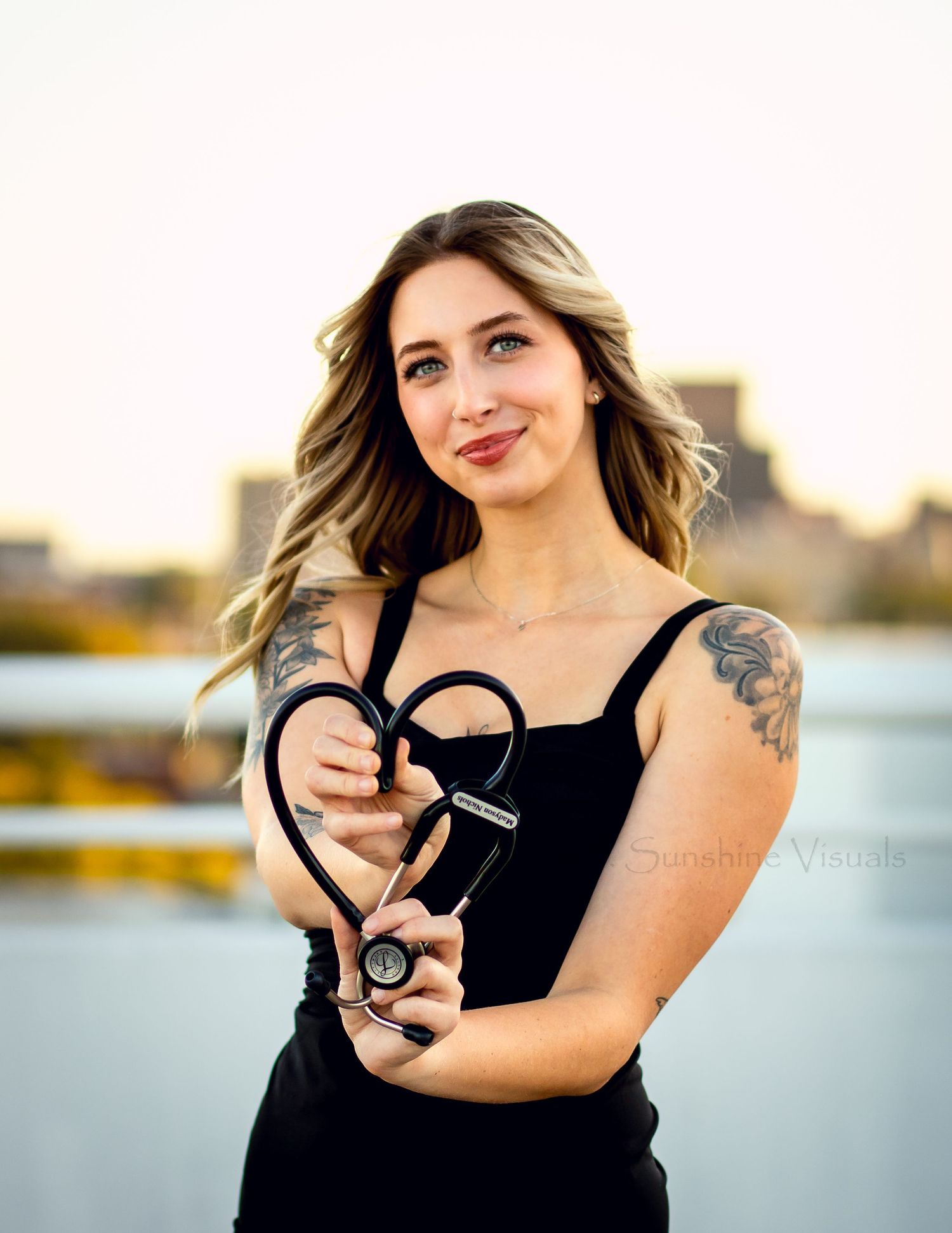 A person in a black top poses outdoors with a heart-shaped stethoscope against a waterfront background.
