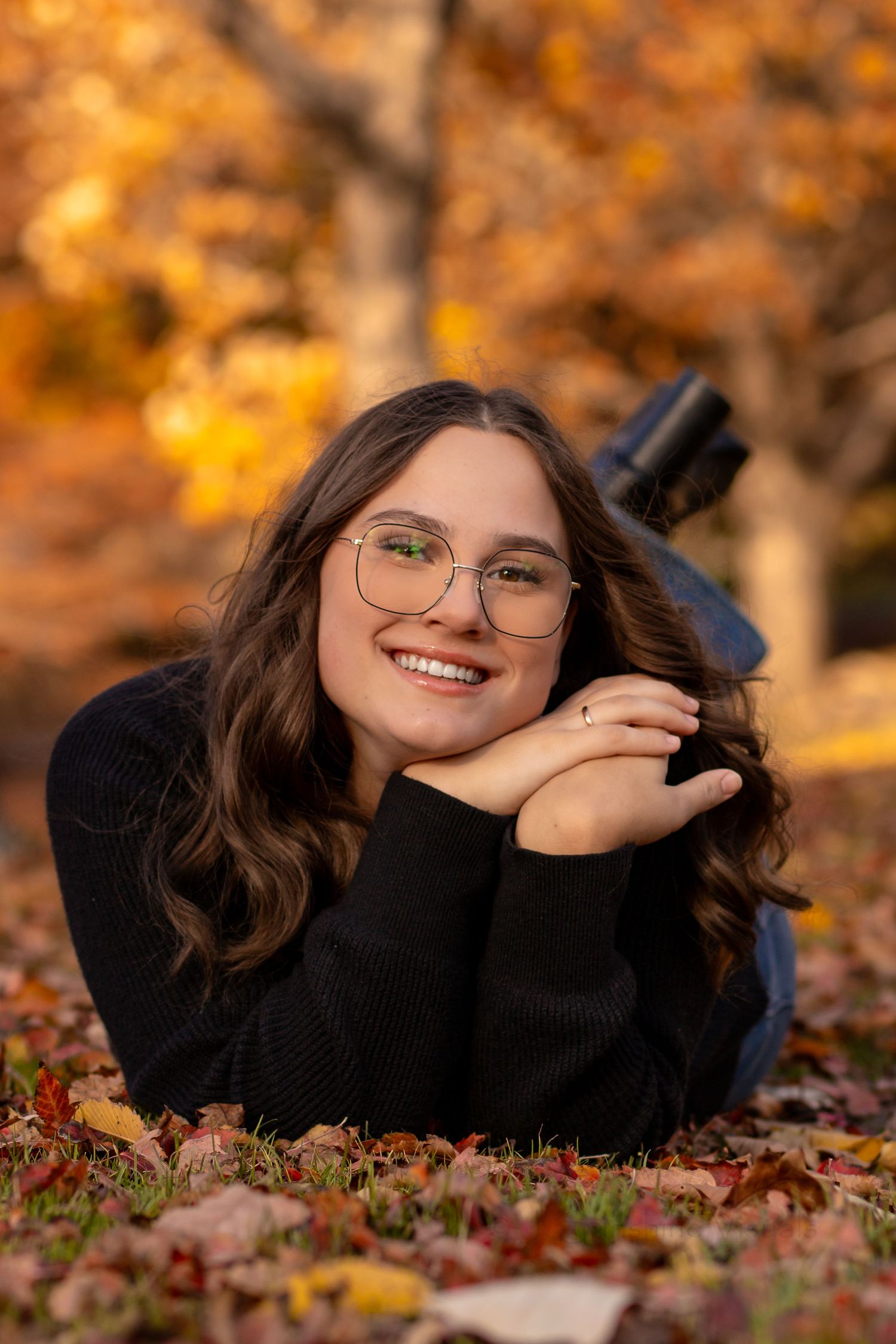 Portrait of someone wearing glasses and a black sweater lying in autumn leaves with golden fall foliage in background.