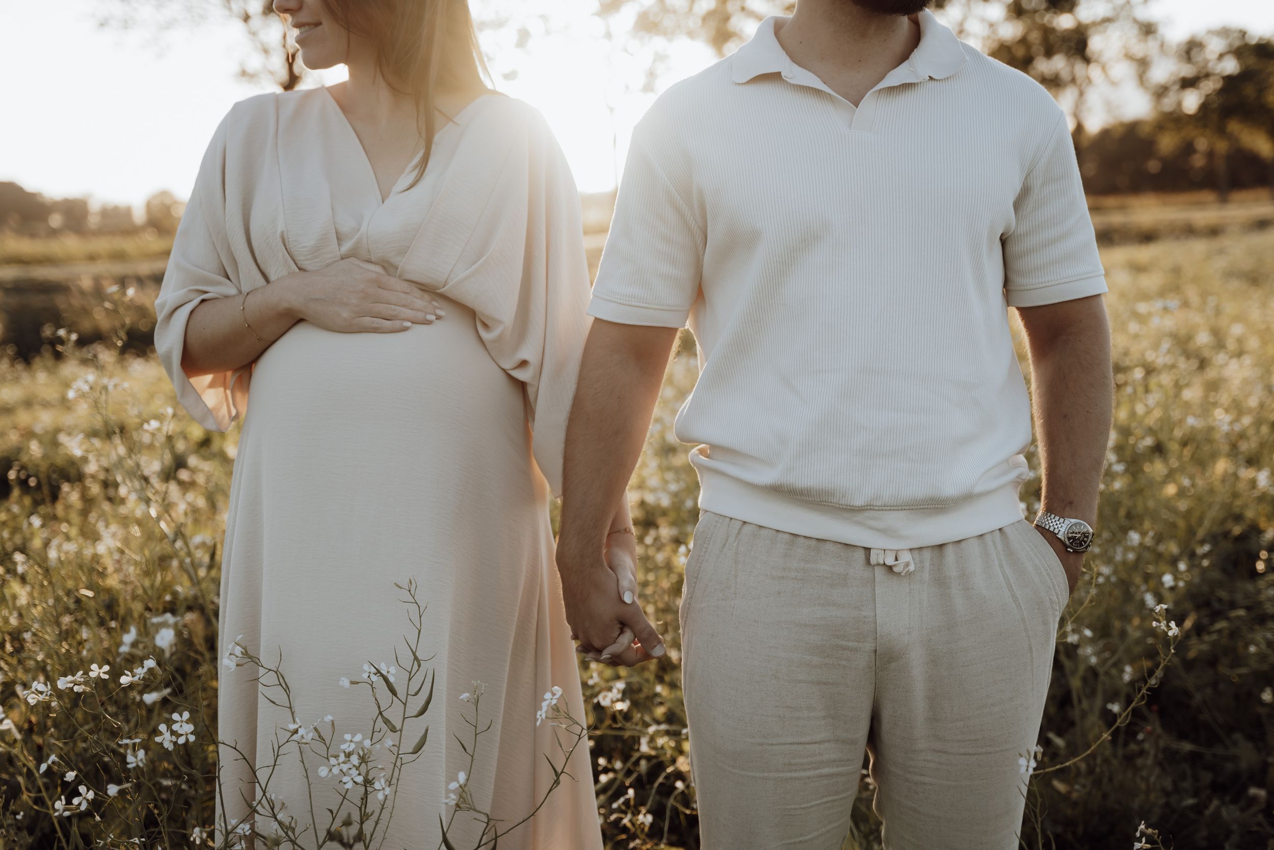 Man en zwangere vrouw staan hand in hand in een veld met hoog gras en witte bloemen bij zonsondergang.
