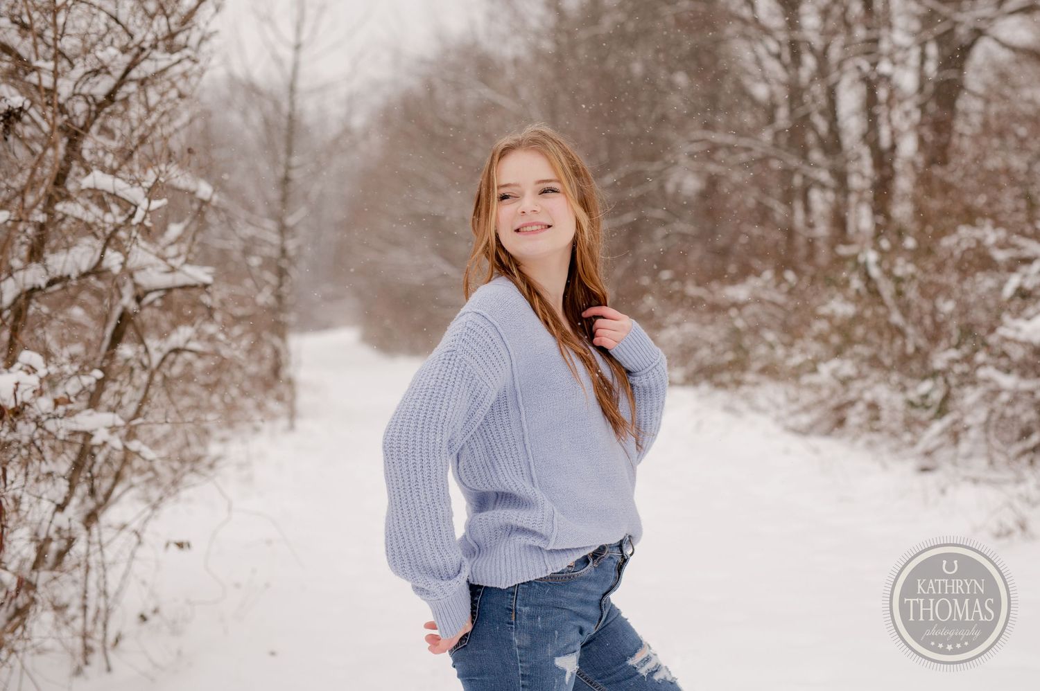 A candid winter portrait featuring someone in a blue knit sweater and jeans on a snowy trail.