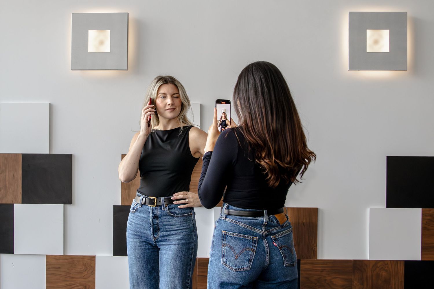 Two women in black tops and blue jeans taking photos of each other against a modern wall with geometric decor.
