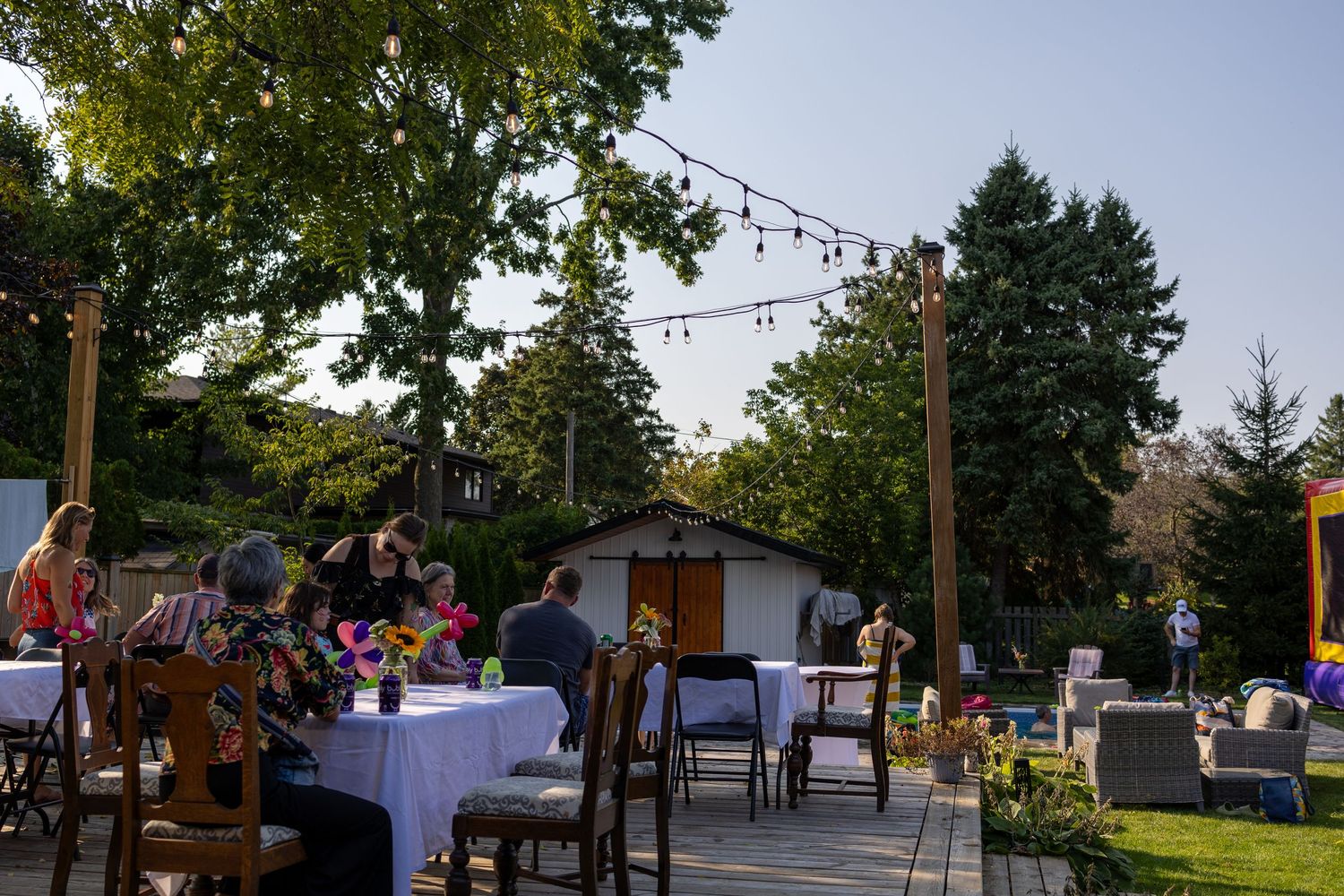An outdoor dining patio at dusk with string lights overhead, featuring elegantly set tables and chairs among garden scenery.