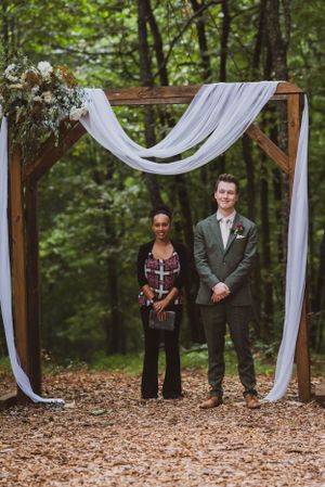 Two people stand under a wooden arch with white fabric draping in a forest setting.