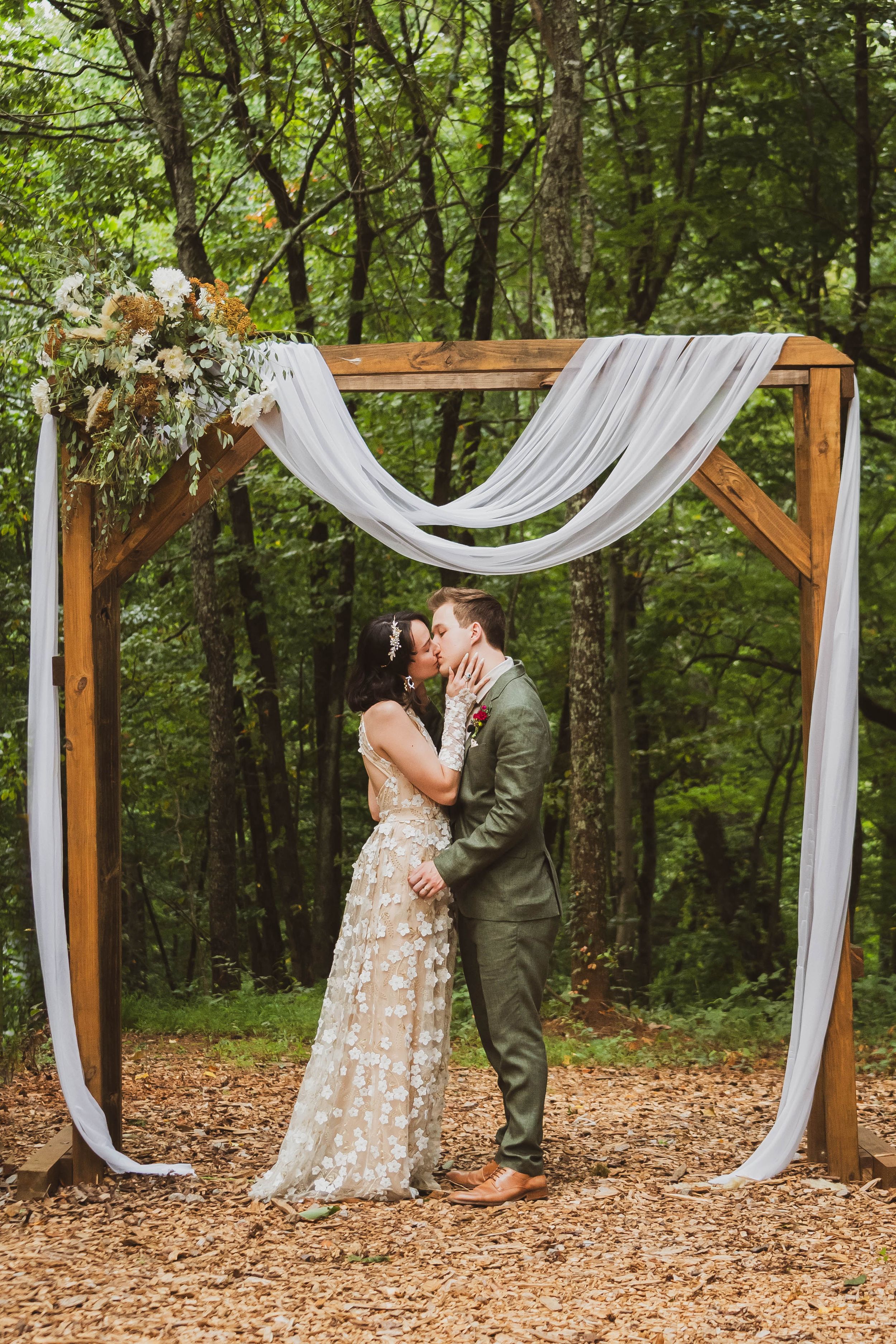 Couple embraces under a wooden wedding arch adorned with flowing white fabric and florals in a forest setting.