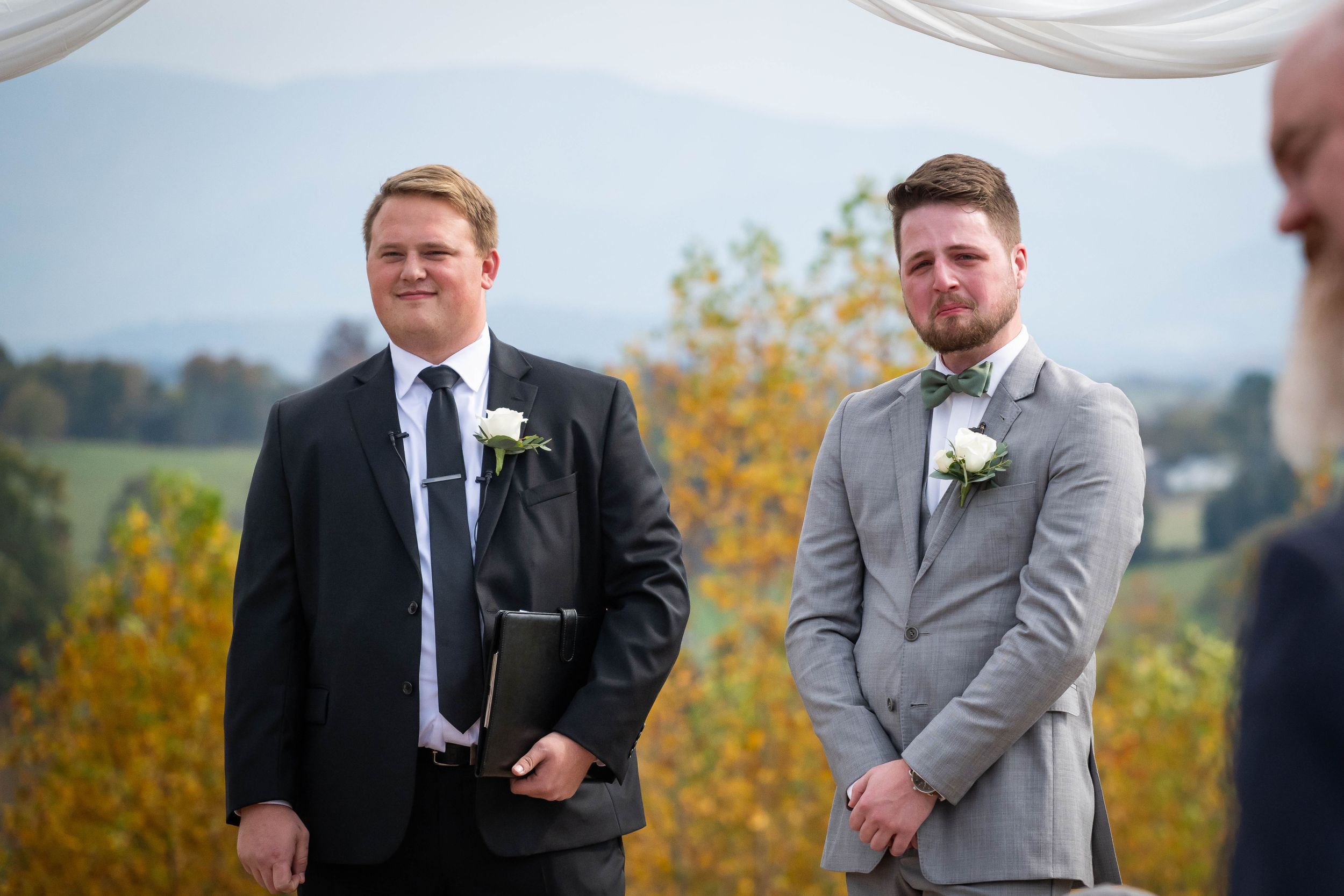 Two suited attendants stand at attention during an outdoor autumn wedding ceremony.