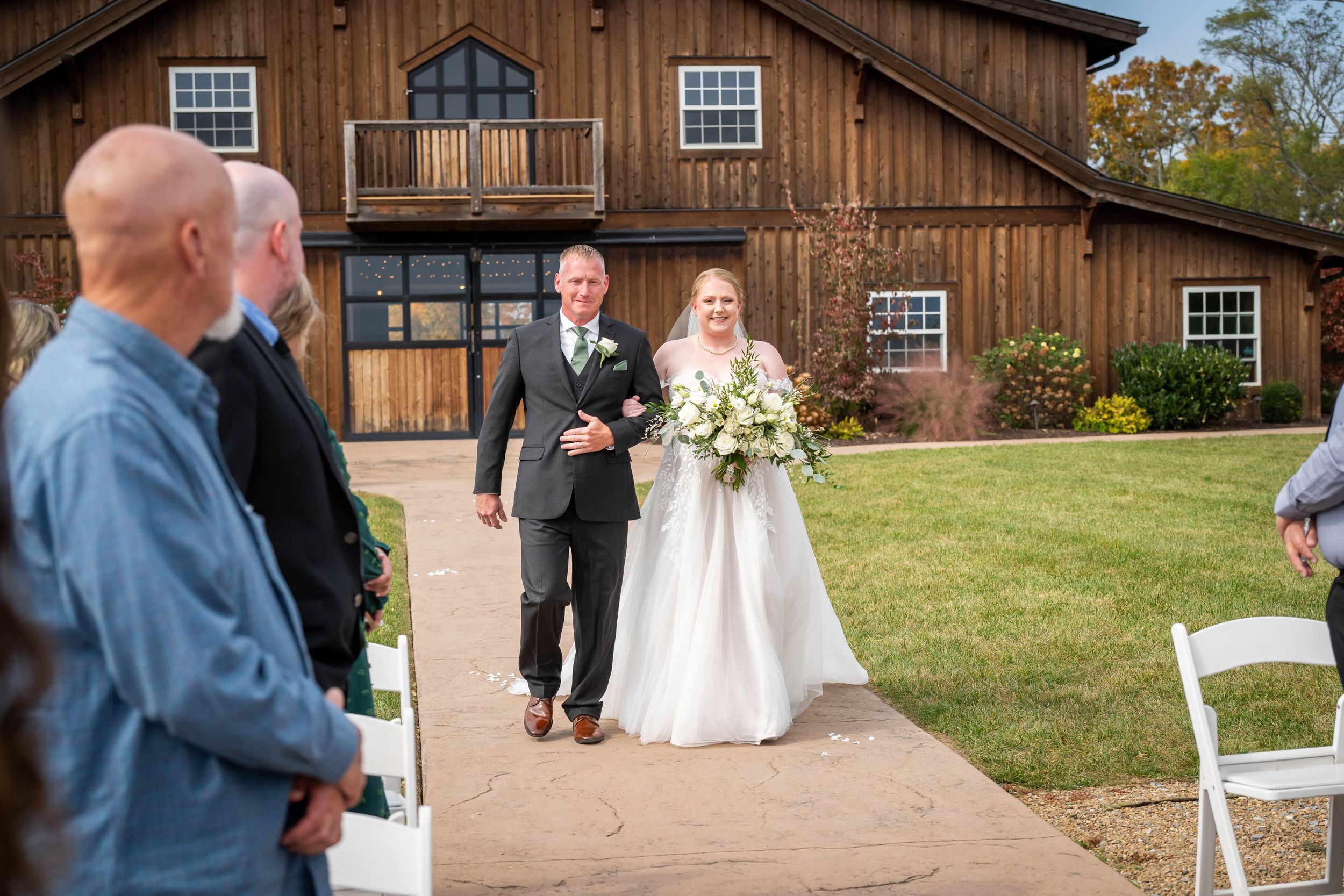 Wedding processional outside a rustic barn venue with wooden architecture and manicured grounds.