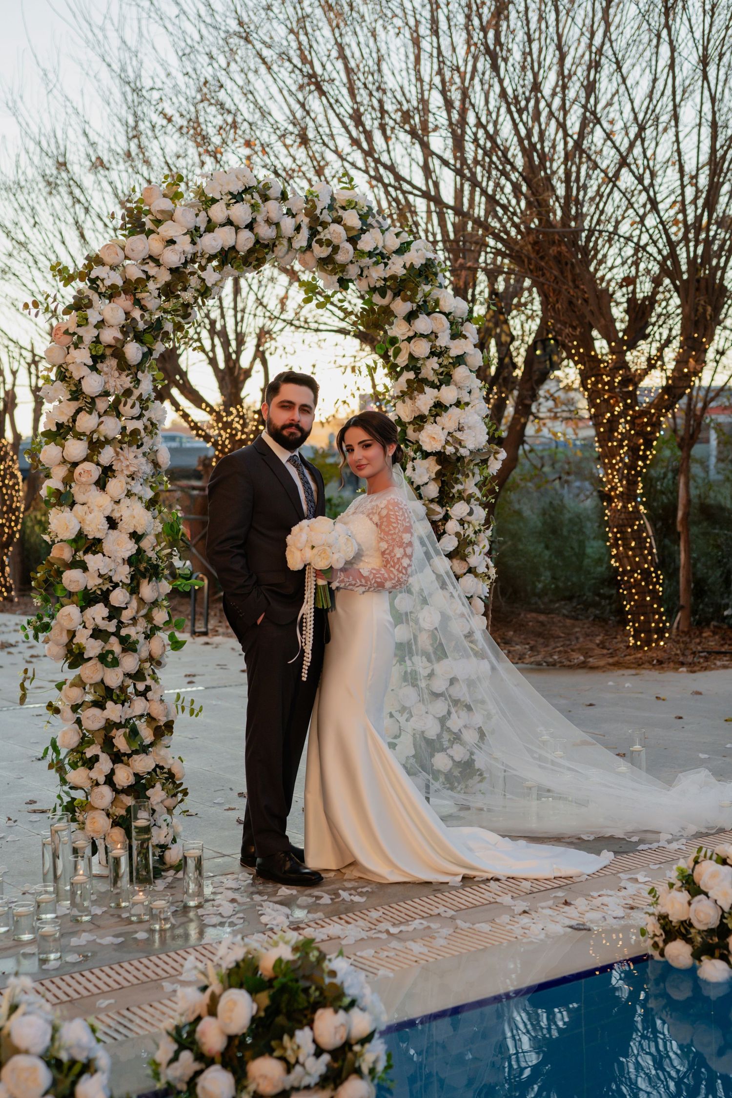A romantic wedding couple poses under a beautiful floral arch with white flowers and twinkle lights by a poolside at dusk.