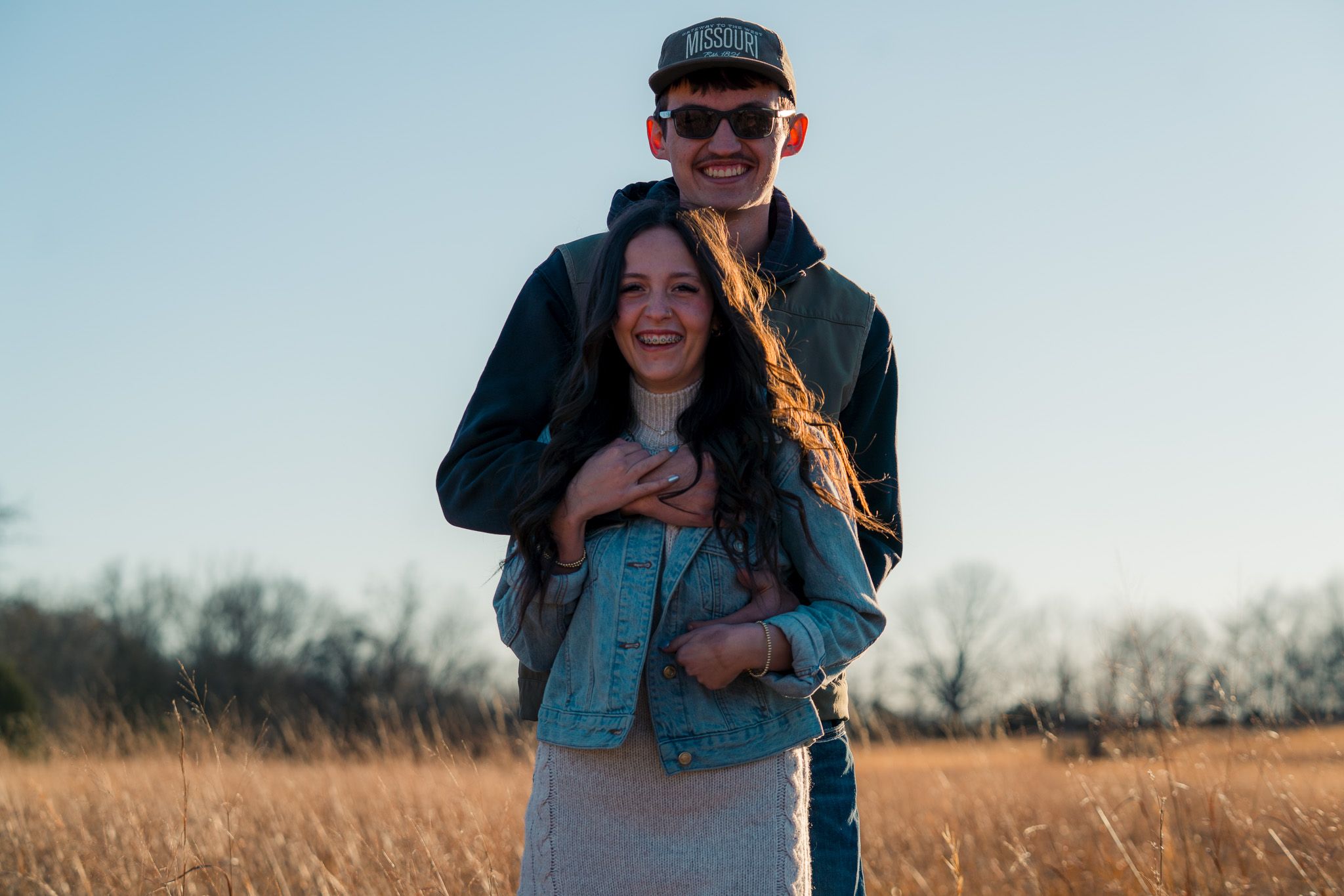 A couple embraces in a golden field during sunset with the person in back wearing a black cap and jacket.
