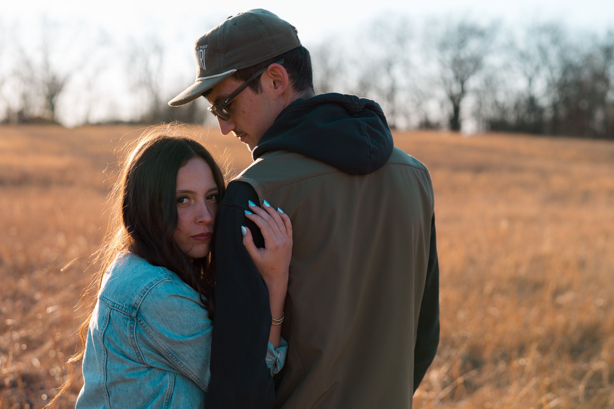 A couple embraces in a golden field during sunset, with the woman wearing a turquoise sweater.